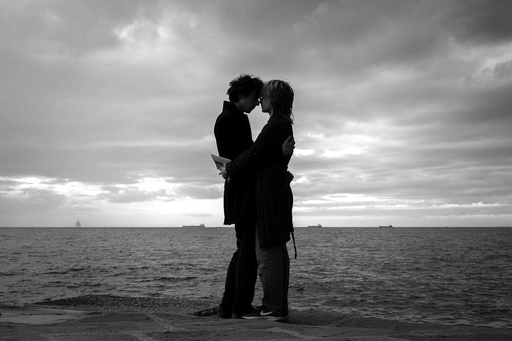 Two people embrace beside the sea beneath a cloudy sky.